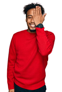 Young african american man with beard wearing casual winter sweater covering one eye with hand, confident smile on face and surprise emotion.