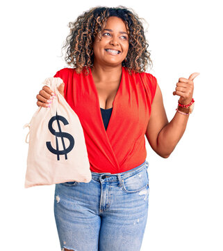 Young african american plus size woman holding money bag with dollar symbol pointing thumb up to the side smiling happy with open mouth