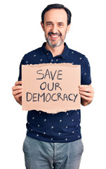 Middle age handsome man holding save our democracy cardboard banner looking positive and happy standing and smiling with a confident smile showing teeth
