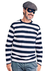 Middle age handsome man wearing burglar mask looking away to side with smile on face, natural expression. laughing confident.