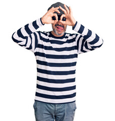 Middle age handsome man wearing burglar mask doing ok gesture like binoculars sticking tongue out, eyes looking through fingers. crazy expression.