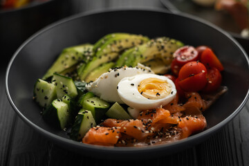 Salad of salmon, cucumber, tomato, egg, avocado and sesame seeds on a black plate, close-up. Vegan restaurant menu