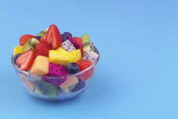 Glass bowl with colorful sliced fruits on blue background. Copy space for text.