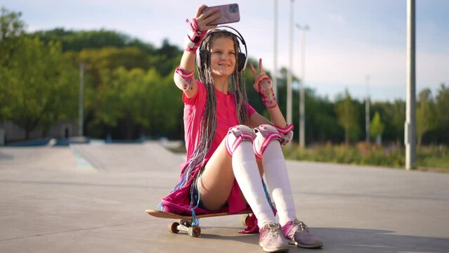 Wide shot portrait of smiling teen girl gesturing peace in slow motion taking selfie sitting on skateboard in sunshine. Happy carefree Caucasian teenage skateboarder photographing with app