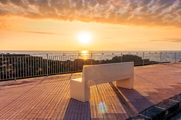 Fototapeta na wymiar beautiful white stone bench on seafront embarkment during beautiful sunrise or sunset with sidewalk with pavement, sea surf and nice cloudy sunset sky