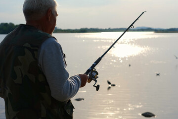 Old man fishing. Senior gray haired fisherman throws a spinning from shoreside at sunset, twisting a coil. Positive elderly male angling at lake, rotating reel rod. Retired fisher enjoying his weekend