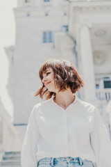 Wind hair style. A portrait of a woman outdoors, her shoulder-length brown hair blowing in the wind. Dressed in a white shirt against a light building.