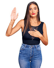 Young hispanic woman wearing casual clothes swearing with hand on chest and open palm, making a...