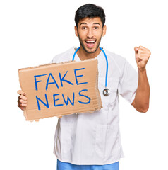 Young handsome man wearing doctor uniform holding fake news banner screaming proud, celebrating...