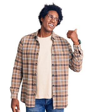 Handsome african american man with afro hair wearing casual clothes and glasses smiling with happy face looking and pointing to the side with thumb up.