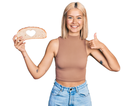 Beautiful young blonde woman holding bread loaf with heart shape smiling happy and positive, thumb up doing excellent and approval sign