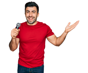 Hispanic man with beard singing song using microphone celebrating victory with happy smile and winner expression with raised hands