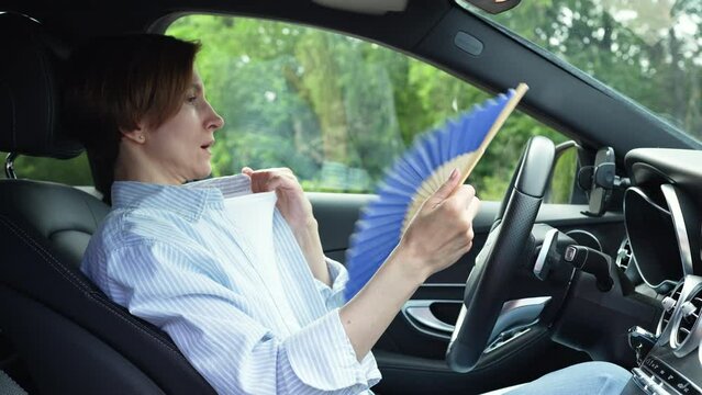 Tired exhausted middle aged woman waving blue fan suffers from stuffiness driving car on summer hot weather. Overheating, sultriness, high temperature, swelter in car with broken air conditioner.