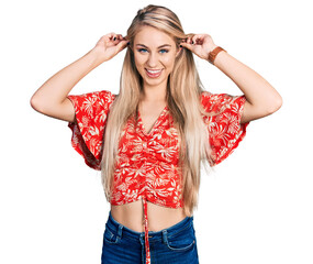 Beautiful young blonde woman wearing summer shirt smiling pulling ears with fingers, funny gesture....