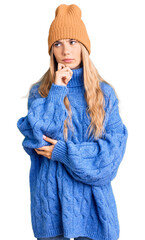 Beautiful caucasian woman with blonde hair wearing wool sweater and winter hat serious face thinking about question with hand on chin, thoughtful about confusing idea