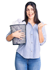 Young beautiful girl holding paper bin full of crumpled papers pointing thumb up to the side smiling happy with open mouth