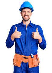 Young handsome man wearing worker uniform and hardhat success sign doing positive gesture with hand, thumbs up smiling and happy. cheerful expression and winner gesture.