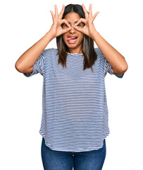 Young latin girl wearing casual clothes doing ok gesture like binoculars sticking tongue out, eyes looking through fingers. crazy expression.