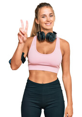 Young blonde woman wearing gym clothes and using headphones showing and pointing up with fingers number two while smiling confident and happy.