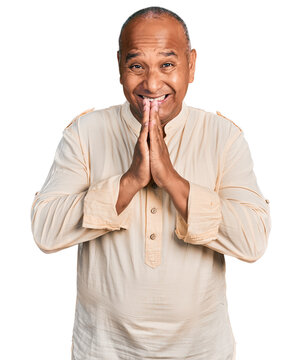 Hispanic middle age man wearing boho shirt praying with hands together asking for forgiveness smiling confident.