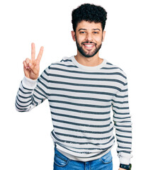 Fototapeta premium Young arab man with beard wearing casual striped sweater smiling looking to the camera showing fingers doing victory sign. number two.