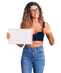 Young hispanic woman with tattoo holding empty white chalkboard pointing finger to one self smiling happy and proud