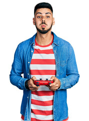 Young hispanic man with beard playing video game holding controller puffing cheeks with funny face. mouth inflated with air, catching air.