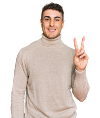 Hispanic young man wearing casual turtleneck sweater smiling with happy face winking at the camera doing victory sign. number two.