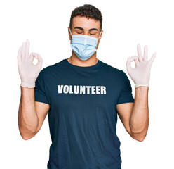 Hispanic young man wearing medical mask and volunteer t shirt relax and smiling with eyes closed doing meditation gesture with fingers. yoga concept.