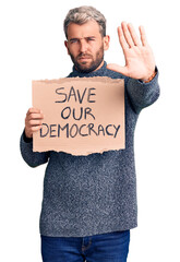 Young blond man holding save our democracy cardboard banner with open hand doing stop sign with serious and confident expression, defense gesture