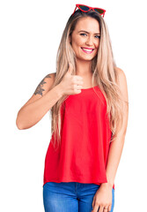 Young beautiful blonde woman wearing sleeveless t-shirt and sunglasses doing happy thumbs up gesture with hand. approving expression looking at the camera showing success.