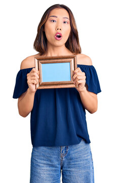 Young beautiful chinese girl holding empty frame scared and amazed with open mouth for surprise, disbelief face