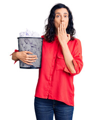 Young beautiful hispanic woman holding paper bin full of crumpled papers covering mouth with hand, shocked and afraid for mistake. surprised expression