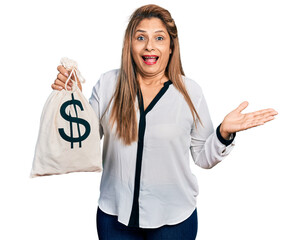 Middle age latin woman holding dollars bag celebrating achievement with happy smile and winner...