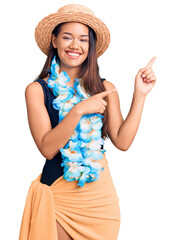 Young beautiful latin girl wearing hawaiian lei and summer hat smiling and looking at the camera pointing with two hands and fingers to the side.