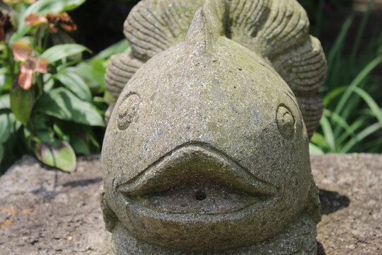 A photo of a stone statue of a fish against a background of green plants and a muddy floor. 