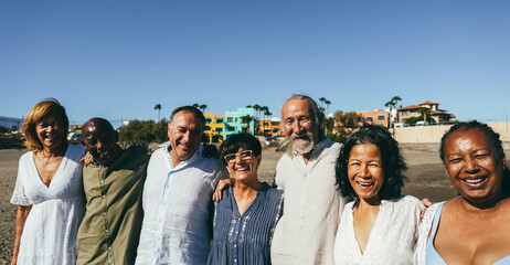Happy senior people having fun walking on the beach at sunset wearing summer clothes - Joyful elderly lifestyle, pensioner vacation and travel concept - Main focus on center friends faces