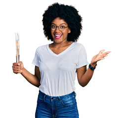 Young african american woman holding paintbrushes celebrating achievement with happy smile and...