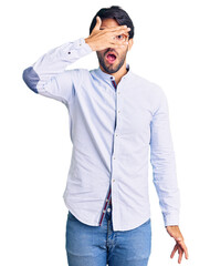 Fototapeta premium Handsome hispanic man wearing business shirt and glasses peeking in shock covering face and eyes with hand, looking through fingers with embarrassed expression.