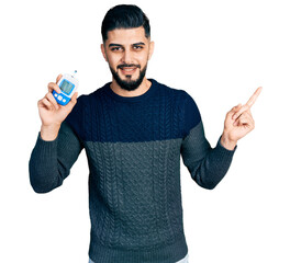 Young arab man with beard holding glucometer device smiling happy pointing with hand and finger to the side