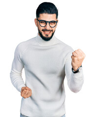 Naklejka premium Young arab man with beard wearing elegant turtleneck sweater and glasses celebrating surprised and amazed for success with arms raised and eyes closed. winner concept.
