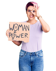 Young beautiful woman with pink hair holding woman power banner smiling happy doing ok sign with hand on eye looking through fingers