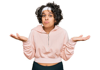 Young hispanic woman with curly hair wearing sportswear clueless and confused expression with arms and hands raised. doubt concept.