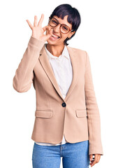 Young brunette woman with short hair wearing business jacket and glasses smiling positive doing ok sign with hand and fingers. successful expression.