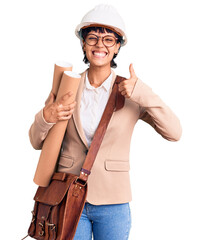 Young brunette woman with short hair wearing safety helmet holding blueprints smiling happy and positive, thumb up doing excellent and approval sign
