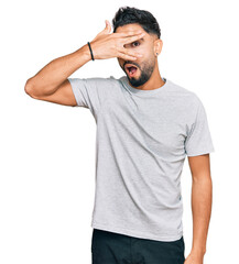 Young man with beard wearing casual grey tshirt peeking in shock covering face and eyes with hand, looking through fingers with embarrassed expression.