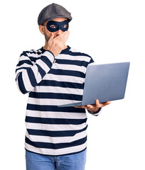 Young handsome man wearing burglar mask using laptop covering mouth with hand, shocked and afraid for mistake. surprised expression