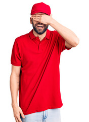 Young handsome man with beard wearing delivery uniform smiling and laughing with hand on face covering eyes for surprise. blind concept.