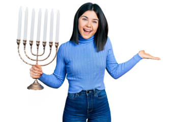 Young brunette woman with blue eyes holding menorah hanukkah jewish candle celebrating achievement with happy smile and winner expression with raised hand
