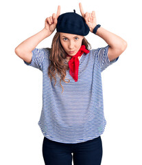 Young beautiful blonde woman wearing french beret and striped t-shirt doing funny gesture with finger over head as bull horns